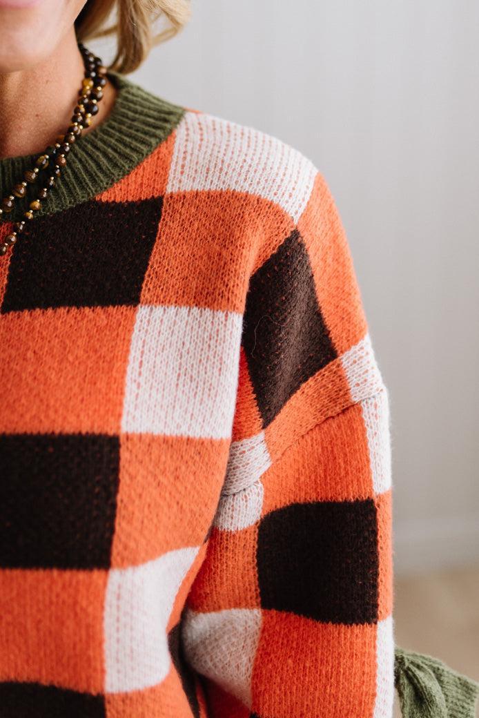 Blonde woman wearing a orange, brown and cream check sweater with olive green ribbed trim and bows at the elbow.