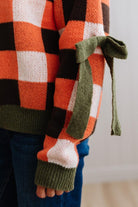Blonde woman wearing a orange, brown and cream check sweater with olive green ribbed trim and bows at the elbow.