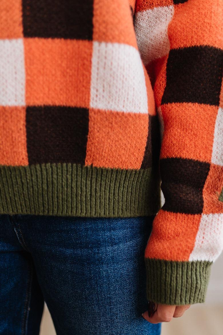 Blonde woman wearing a orange, brown and cream check sweater with olive green ribbed trim and bows at the elbow.
