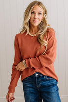 A woman wearing a rust-orange long-sleeve knit sweater and blue jeans, standing against a white paneled wall.