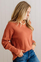 A woman wearing a long-sleeve knit sweater in a muted terracotta or burnt orange color, paired with blue jeans and a pearl necklace, standing against a white paneled wall.