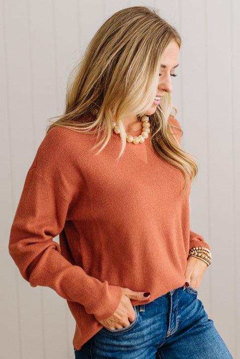 A woman wearing a long-sleeve knit sweater in a muted terracotta or burnt orange color, paired with blue jeans and a pearl necklace, standing against a white paneled wall.