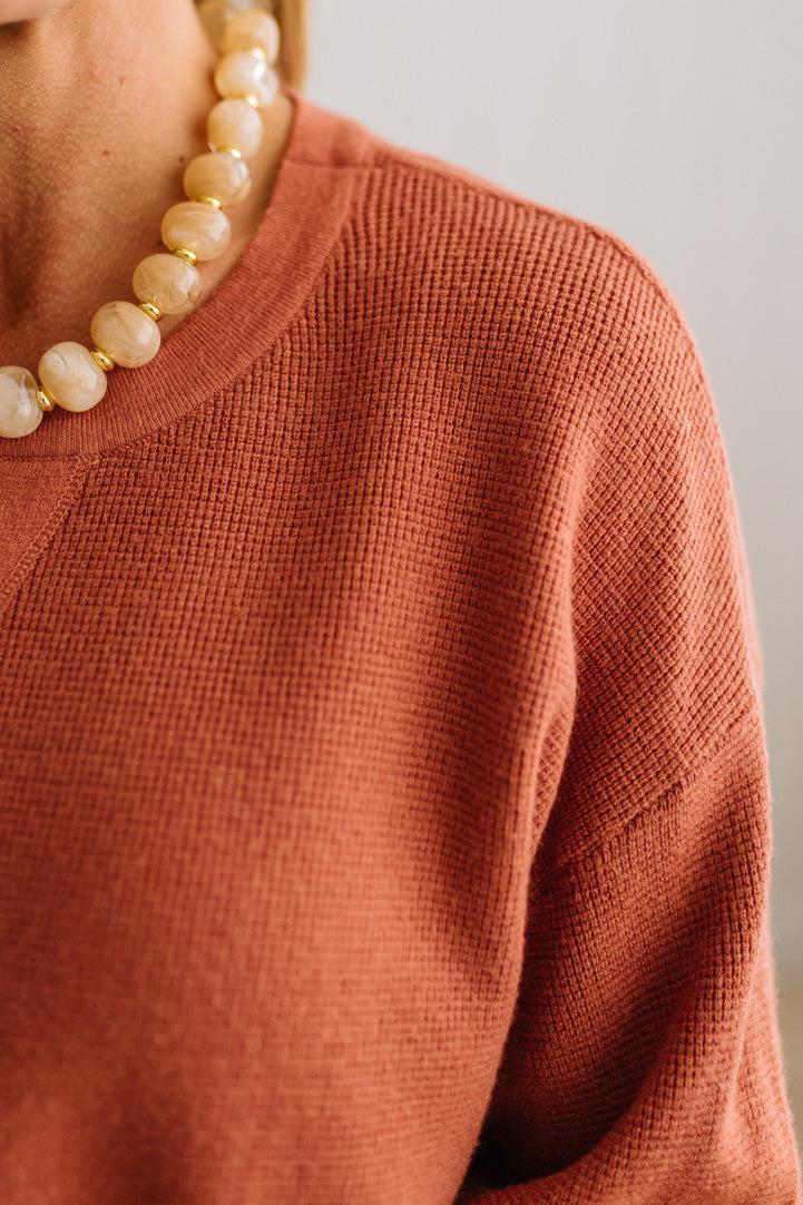 Close-up of a person wearing a textured terracotta orange knit top and a chunky cream beaded necklace with gold spacer rings, against a neutral light gray background.