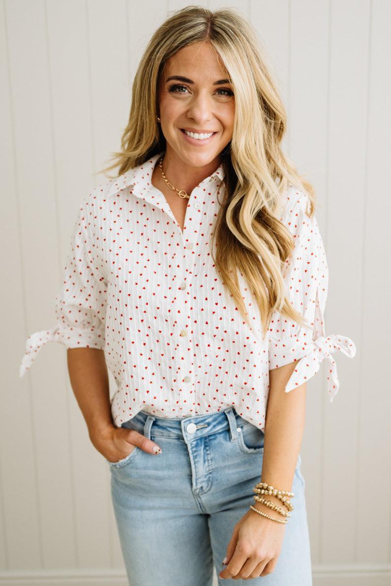 Woman wearing a white blouse with red mini heart patterns and light blue jeans against a neutral background