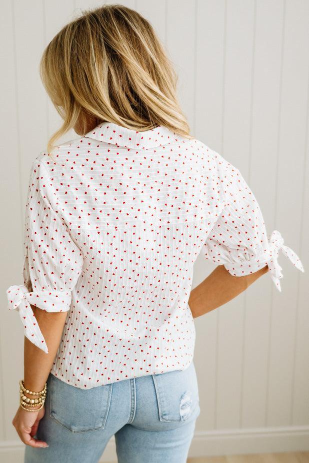 Woman wearing a white blouse with red mini heart patterns and light blue jeans against a neutral background