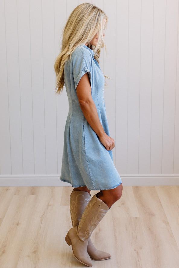 Woman wearing a light blue dress and brown boots against a white wall.