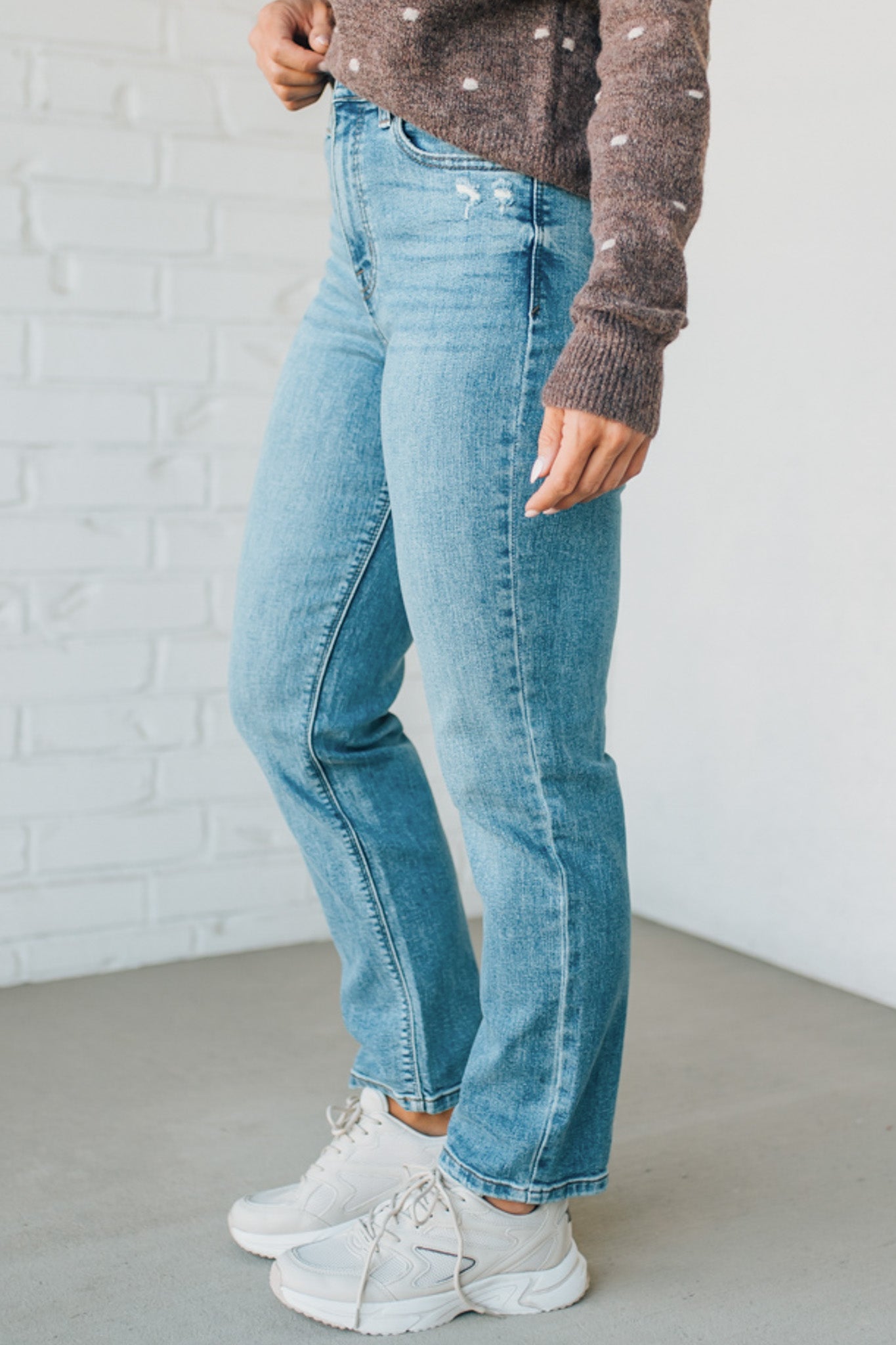 Person wearing blue jeans and a brown sweater against a white brick wall.