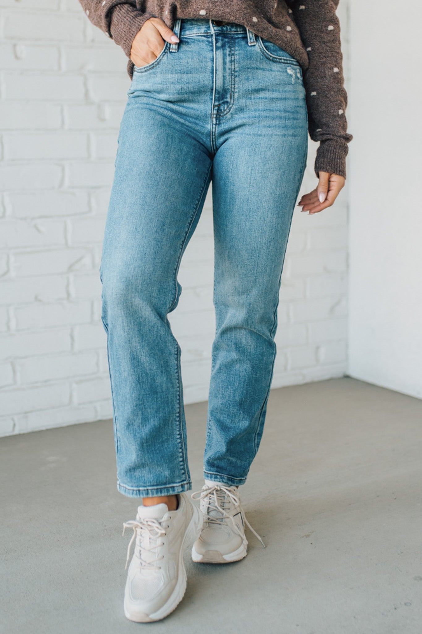 Person wearing blue jeans and white sneakers against a white brick wall.