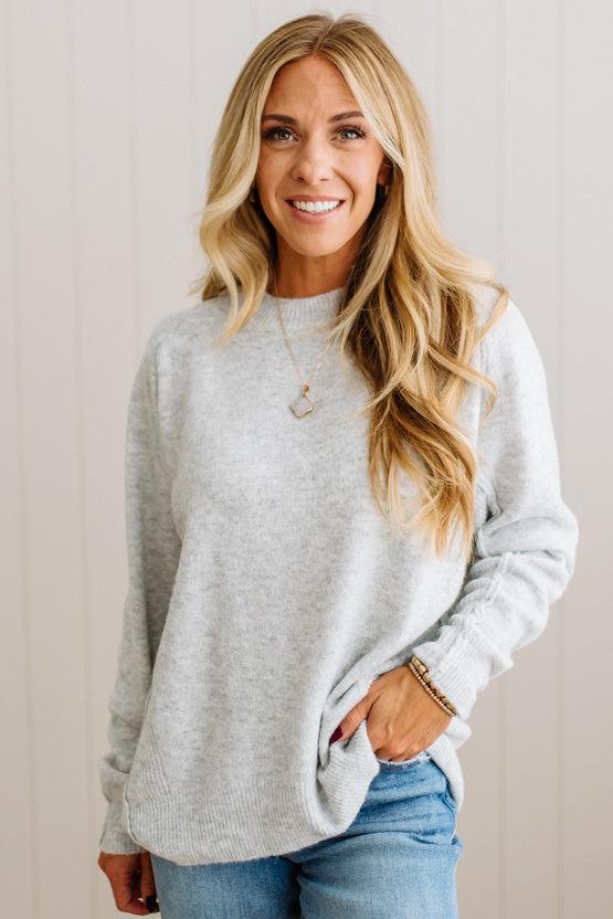 BLONDE WOMAN WEARING A HIGH-LOW HEM, LONG SLEEVE, SUPER SOFT, OVERSIZED CREW NECK SWEATER WITH RIB DETAILS AND STANDING IN A STUDIO WITH LIGHT WOOD FLOOR AND PANELED WALL BEHIND.
