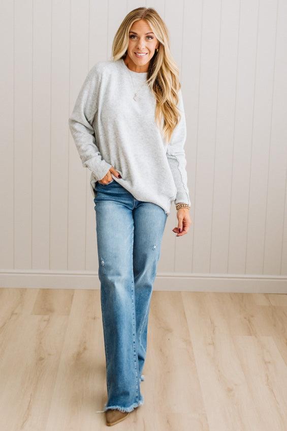 BLONDE WOMAN WEARING A HIGH-LOW HEM, LONG SLEEVE, SUPER SOFT, OVERSIZED CREW NECK SWEATER WITH RIB DETAILS AND STANDING IN A STUDIO WITH LIGHT WOOD FLOOR AND PANELED WALL BEHIND.