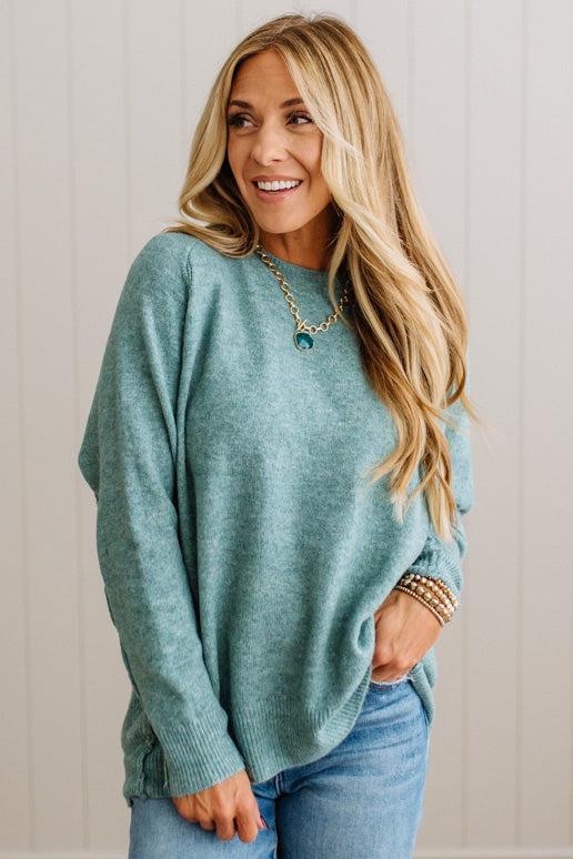 BLONDE WOMAN WEARING A HIGH-LOW HEM, LONG SLEEVE, SUPER SOFT, OVERSIZED CREW NECK SWEATER WITH RIB DETAILS AND STANDING IN A STUDIO WITH LIGHT WOOD FLOOR AND PANELED WALL BEHIND.