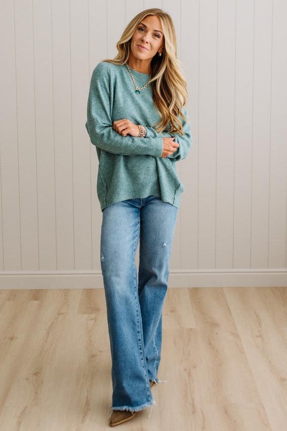 BLONDE WOMAN WEARING A HIGH-LOW HEM, LONG SLEEVE, SUPER SOFT, OVERSIZED CREW NECK SWEATER WITH RIB DETAILS AND STANDING IN A STUDIO WITH LIGHT WOOD FLOOR AND PANELED WALL BEHIND.
