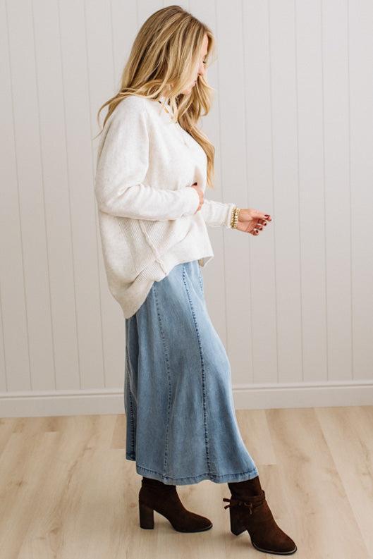 BLONDE WOMAN WEARING A HIGH-LOW HEM, LONG SLEEVE, SUPER SOFT, OVERSIZED CREW NECK SWEATER WITH RIB DETAILS AND STANDING IN A STUDIO WITH LIGHT WOOD FLOOR AND PANELED WALL BEHIND.
