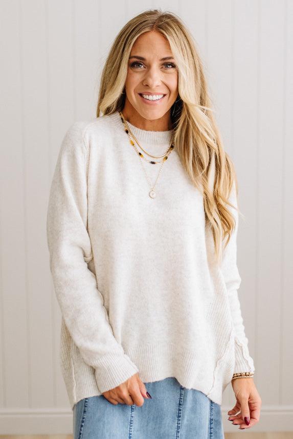 BLONDE WOMAN WEARING A HIGH-LOW HEM, LONG SLEEVE, SUPER SOFT, OVERSIZED CREW NECK SWEATER WITH RIB DETAILS AND STANDING IN A STUDIO WITH LIGHT WOOD FLOOR AND PANELED WALL BEHIND.