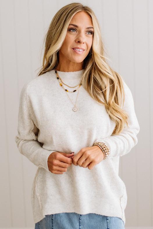 BLONDE WOMAN WEARING A HIGH-LOW HEM, LONG SLEEVE, SUPER SOFT, OVERSIZED CREW NECK SWEATER WITH RIB DETAILS AND STANDING IN A STUDIO WITH LIGHT WOOD FLOOR AND PANELED WALL BEHIND.