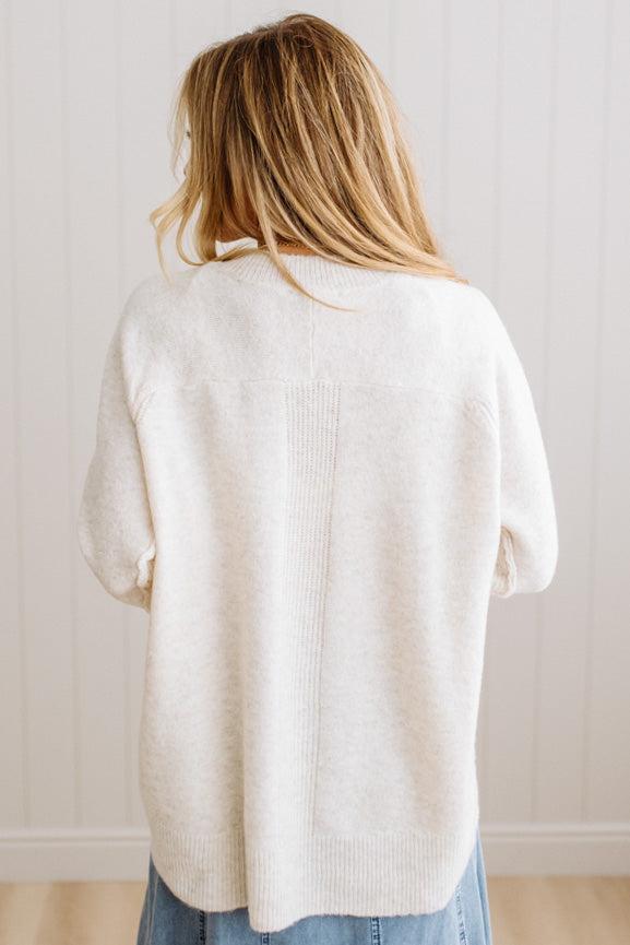 BLONDE WOMAN WEARING A HIGH-LOW HEM, LONG SLEEVE, SUPER SOFT, OVERSIZED CREW NECK SWEATER WITH RIB DETAILS AND STANDING IN A STUDIO WITH LIGHT WOOD FLOOR AND PANELED WALL BEHIND.
