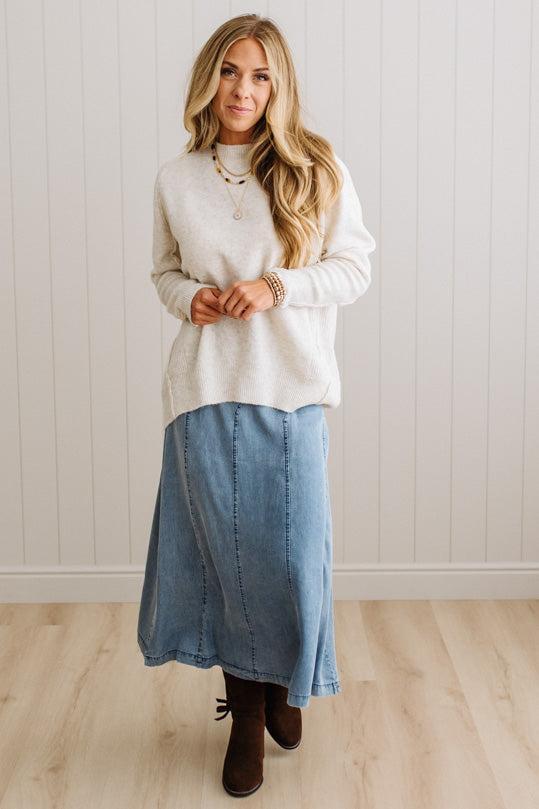 BLONDE WOMAN WEARING A HIGH-LOW HEM, LONG SLEEVE, SUPER SOFT, OVERSIZED CREW NECK SWEATER WITH RIB DETAILS AND STANDING IN A STUDIO WITH LIGHT WOOD FLOOR AND PANELED WALL BEHIND.