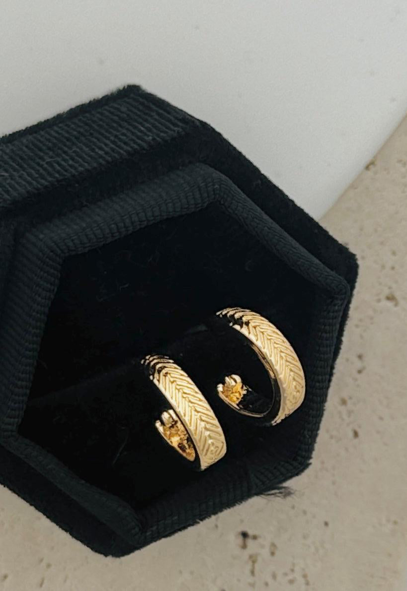 Two gold earrings in a black jewelry box on a light gray surface