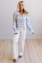A woman modeling a light blue and white plaid long-sleeve blouse with ruffled cuffs and a smocked bodice, paired with white wide-leg pants and brown platform sandals, standing in front of a white paneled wall and light wood floor.