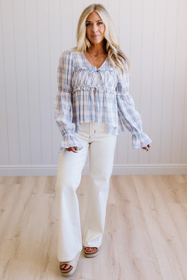 A woman modeling a light blue and white plaid long-sleeve blouse with ruffled cuffs and a smocked bodice, paired with white wide-leg pants and brown platform sandals, standing in front of a white paneled wall and light wood floor.
