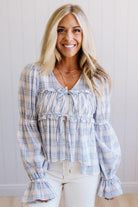 A smiling blonde woman wearing a light blue and white plaid long-sleeve blouse with a ruffled tie neckline and flared cuffs, paired with white pants, standing against a white paneled wall.