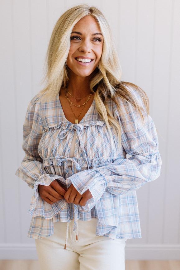 A smiling blonde woman wearing a light blue and white plaid long-sleeve blouse with a ruffled tie neckline and gathered waist, paired with cream pants, standing against a white paneled wall.