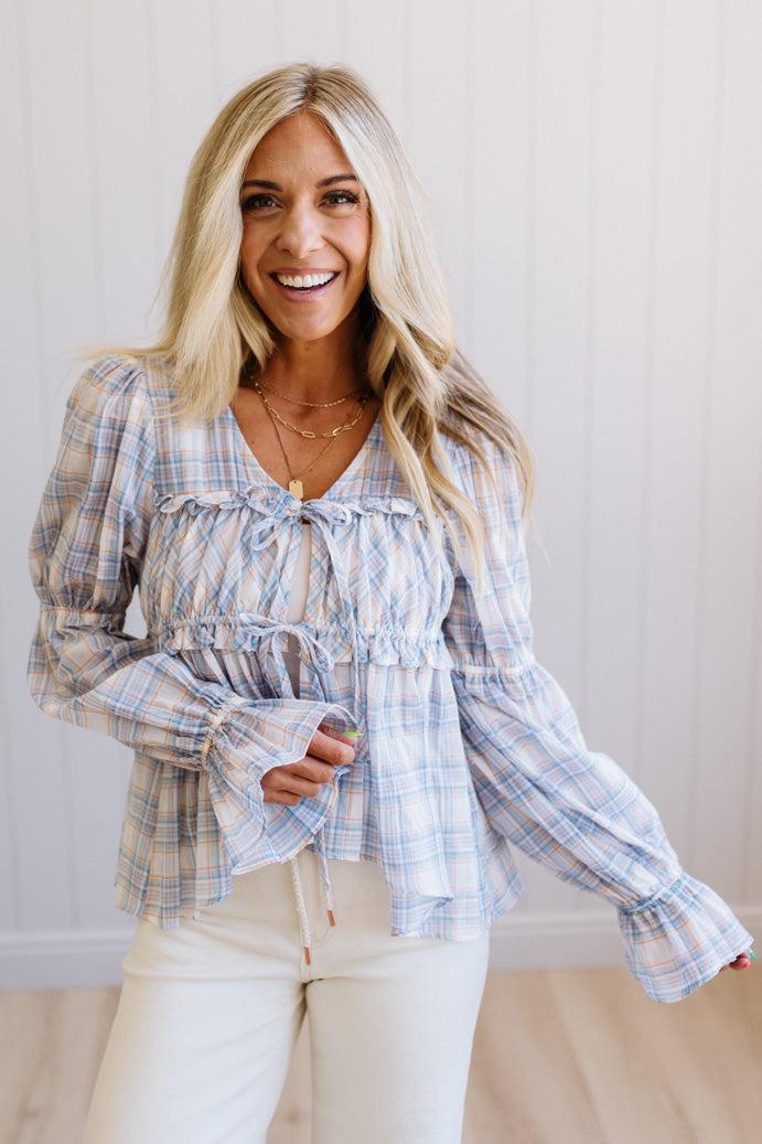 A smiling blonde woman wearing a light blue and white plaid long-sleeve blouse with ruffles and tie details, paired with white pants, standing against a white paneled wall.