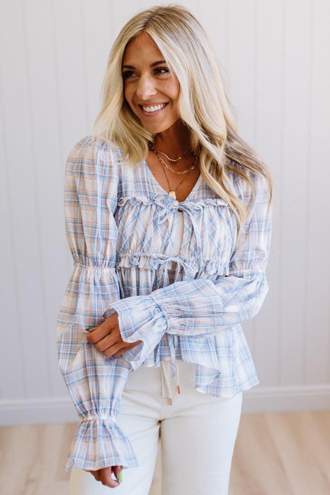 A woman wearing a light blue and white plaid long-sleeve blouse with a ruffled tie neckline and flared cuffs, paired with white pants, standing against a white paneled wall.