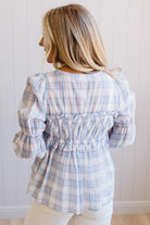 A woman shown from the back wearing a light blue and white plaid long-sleeve blouse with a smocked back detail, paired with cream pants, set against a white paneled wall.