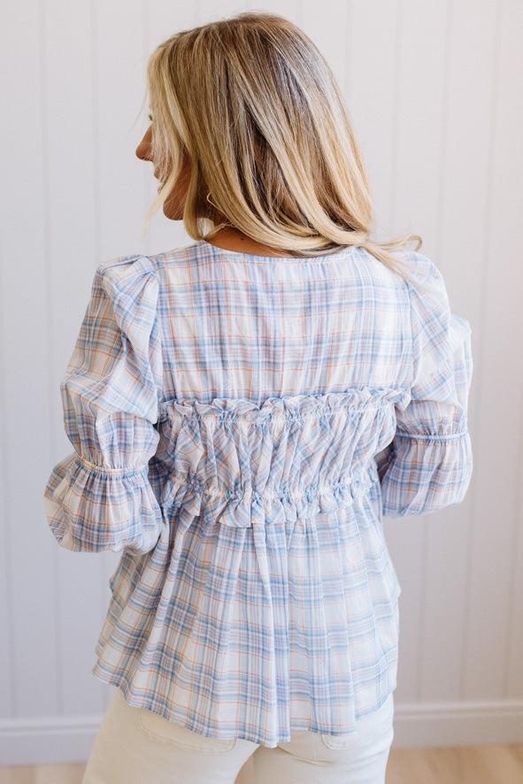 A woman shown from the back wearing a light blue and white plaid long-sleeve blouse with a smocked back detail, paired with cream pants, set against a white paneled wall.