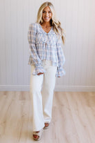 A woman modeling a light blue and white plaid long-sleeve blouse with ruffled cuffs and a tie detail at the neckline, paired with white pants and brown sandals, standing in front of a white paneled wall and light wood floor.