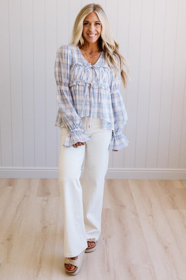A woman modeling a light blue and white plaid long-sleeve blouse with ruffled cuffs and a tie detail at the neckline, paired with white pants and brown sandals, standing in front of a white paneled wall and light wood floor.