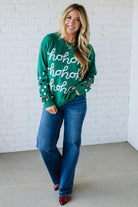 Woman wearing a green sweater with 'ho ho ho' text and blue jeans against a white brick wall.