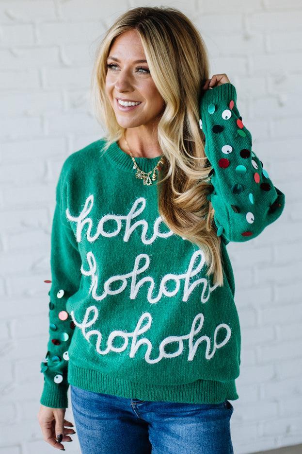Woman wearing a green sweater with 'ho ho ho' text and colorful buttons against a white brick wall.
