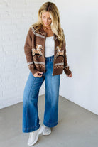 Woman wearing a brown patterned cardigan, white top, and blue jeans against a white brick wall.