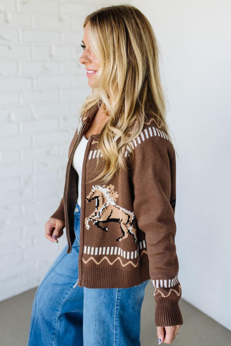 Woman wearing a brown cardigan with horse design against a white brick wall.