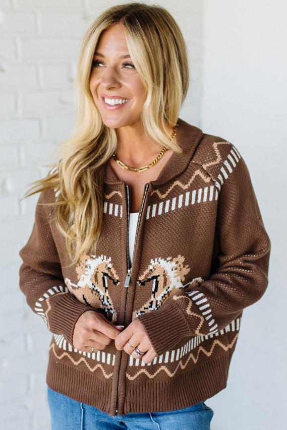 Woman wearing a brown zip-up sweater with a pattern against a white brick wall.