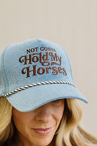 Blonde woman in a brown top showing a denim hat with a brown and white rope across the front, and brown embroidery that says "Not Gonna Hold My Horses"