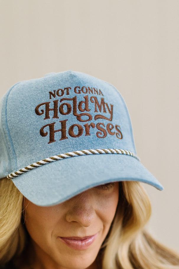 Blonde woman in a brown top showing a denim hat with a brown and white rope across the front, and brown embroidery that says "Not Gonna Hold My Horses"