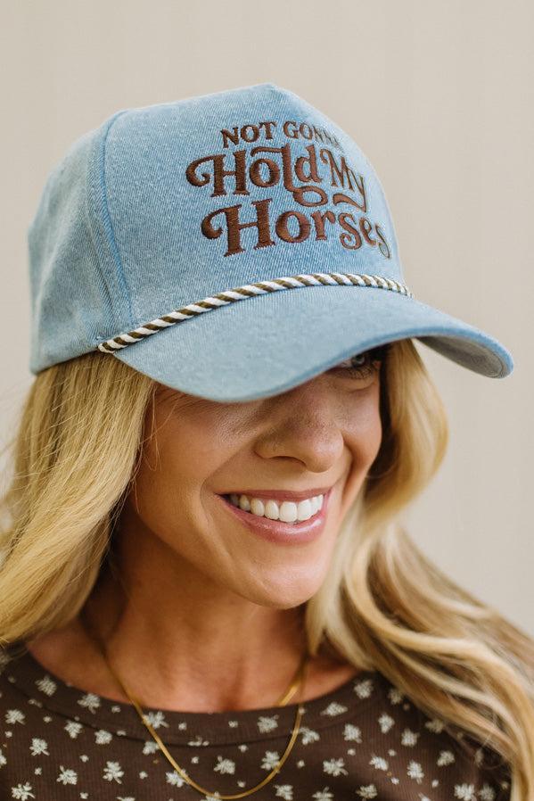 Blonde woman in a brown top showing a denim hat with a brown and white rope across the front, and brown embroidery that says "Not Gonna Hold My Horses"