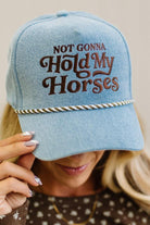 Blonde woman in a brown top showing a denim hat with a brown and white rope across the front, and brown embroidery that says "Not Gonna Hold My Horses"