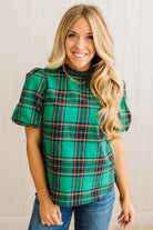Woman wearing a green plaid top with ruffled sleeves against a white wall.