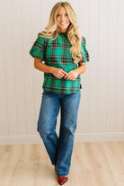 Woman wearing a green plaid top and blue jeans standing against a white paneled wall.