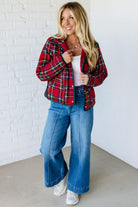 Woman wearing a red plaid jacket, white top, and blue jeans against a white brick wall.