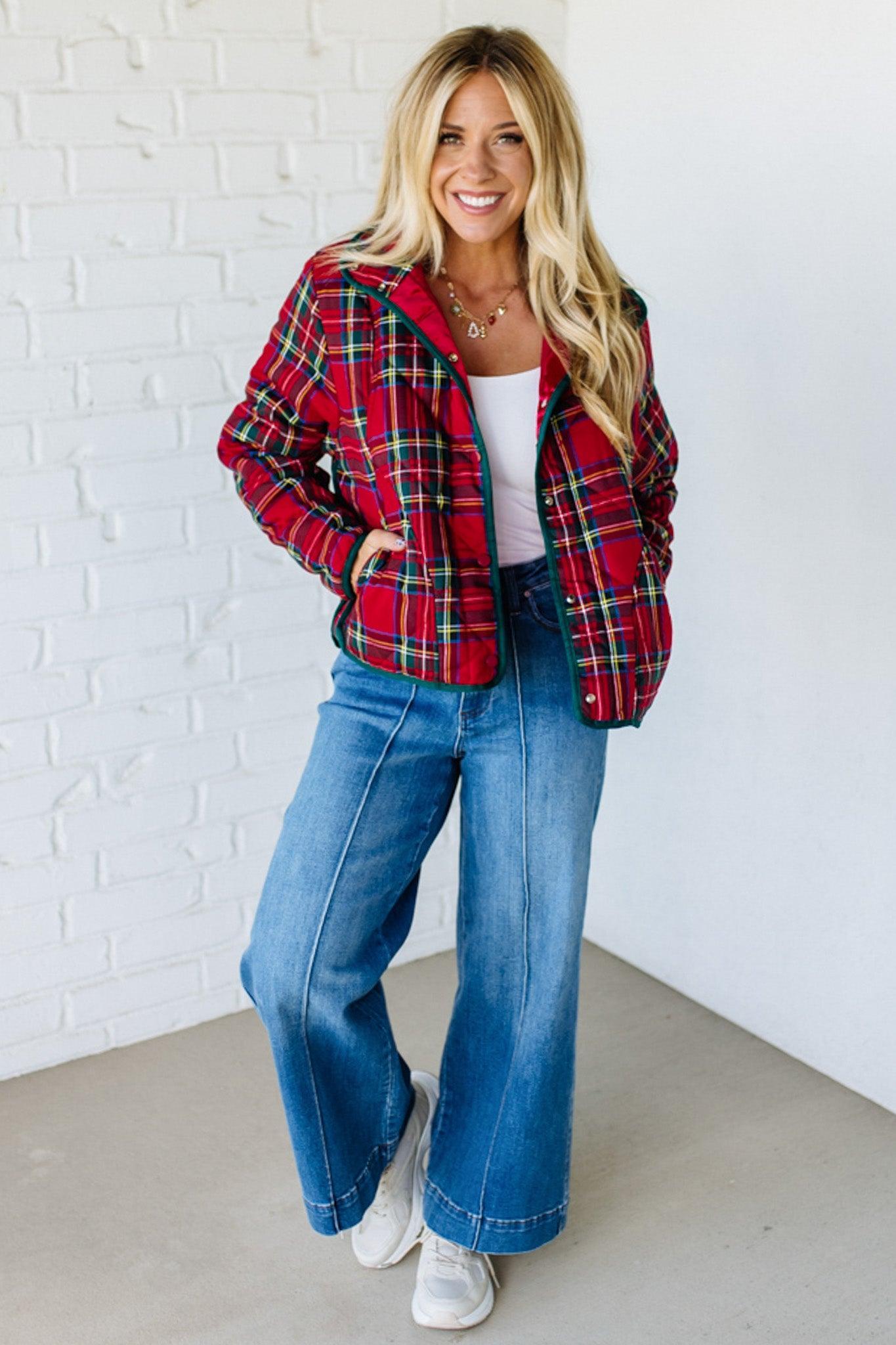 Woman wearing a red plaid jacket, white top, and blue jeans against a white brick wall.