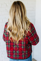 Person wearing a red plaid shirt with long blonde hair, standing against a white brick wall.