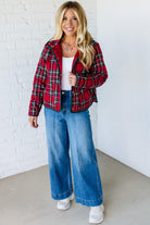 Woman wearing a red plaid shirt, white top, blue jeans, and white sneakers against a white brick wall.