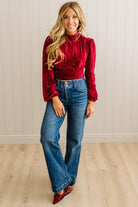 Woman wearing a red blouse and blue jeans standing against a light wooden paneled wall.