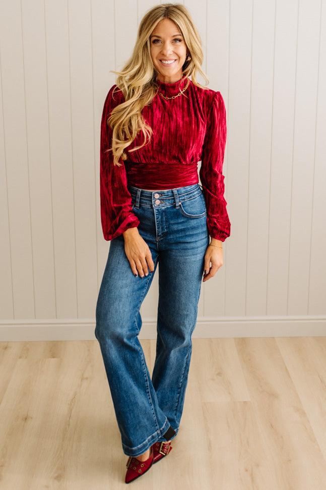 Woman wearing a red blouse and blue jeans standing against a light wooden paneled wall.