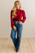 Woman wearing a red blouse and blue jeans standing against a white paneled wall.